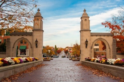 A scenic view of a stone archway leading to a tree-lined street, adorned with autumn foliage and colorful flowers.