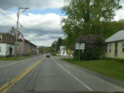 A rural road with a sign for routes 27 and 234, lined with trees and buildings under a cloudy sky.
