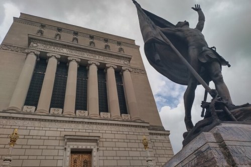 A statue of a soldier holding a flag stands in front of the Indiana World War Memorial, with cloudy skies above.