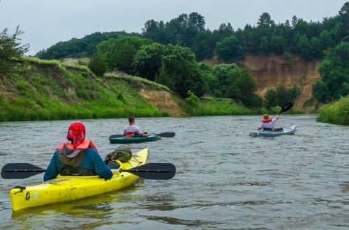 Three people kayaking on a calm river surrounded by lush greenery and steep, sandy banks under a cloudy sky.