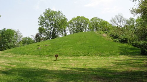 A grassy burial mound surrounded by trees, with a small sign in the foreground indicating its historical significance.