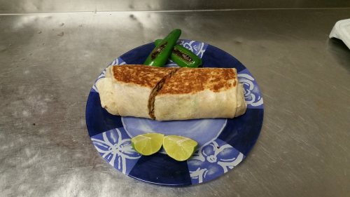 A grilled burrito on a blue plate, accompanied by two lime wedges and two green jalapeños.