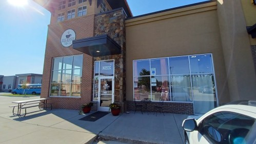 Exterior view of a coffee shop with a stone facade, large windows, and outdoor seating area. Bright sunny day.