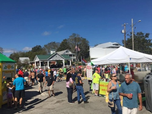 A bustling outdoor festival with crowds, food stalls, and a stage in the background under a clear blue sky.
