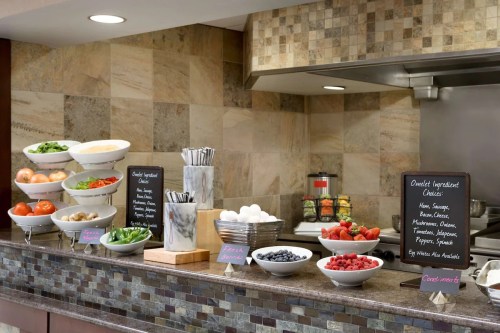 A buffet station with bowls of fresh vegetables, fruits, and condiments, alongside chalkboard menus and utensils.
