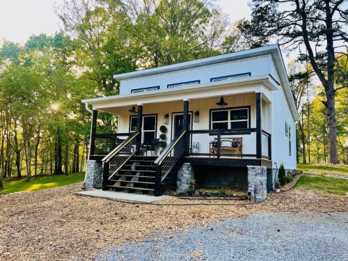 A modern two-story house with a porch, surrounded by trees and gravel driveway, bathed in warm sunlight.
