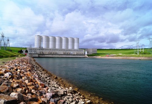 A large industrial facility with silos beside a calm river, surrounded by green fields and power lines under a cloudy sky.