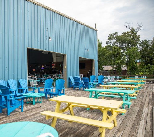 Outdoor seating area with colorful chairs and tables on a wooden deck, under a blue metal building.
