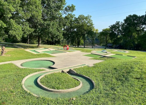A mini-golf course with green turf and concrete paths, surrounded by trees and a view of a river in the background.