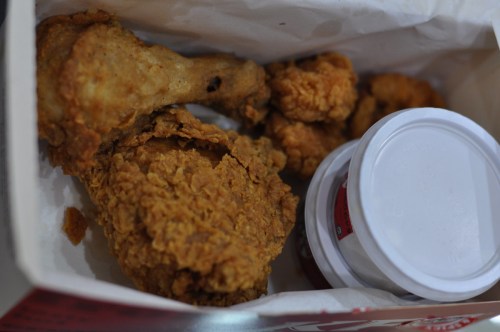 A takeout box containing crispy fried chicken pieces and two small containers of dipping sauce.
