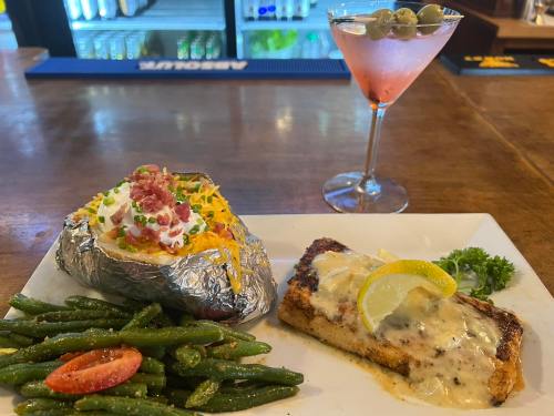 A plate with grilled fish, a loaded baked potato, green beans, and a martini with olives in a bar setting.