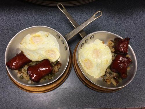 Two small metal pans with fried eggs and sausage links on a bed of hash browns, placed on wooden trivets.