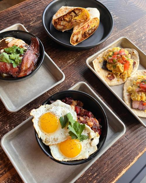 A spread of breakfast dishes including eggs, bacon, and wraps on wooden and metal trays.