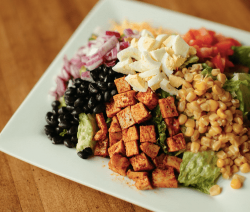 A colorful salad with diced tofu, black beans, corn, lettuce, tomatoes, red onion, and hard-boiled eggs on a white plate.