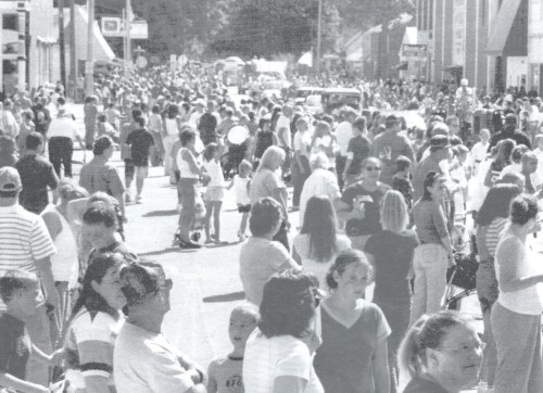 A crowded street filled with people enjoying a community event, with vendors and activities in the background.
