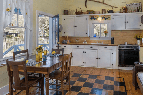 Cozy kitchen with white cabinets, wooden floors, a dining table, and sunflowers, featuring a window and rustic decor.
