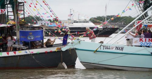 Colorful boats decorated with flags and banners are docked in a harbor, with people celebrating on board.