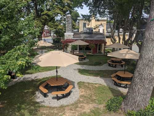 A landscaped outdoor seating area with picnic tables and umbrellas surrounded by trees and gravel pathways.