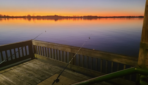 A serene lake at sunset, with fishing rods set up on a wooden dock, reflecting vibrant orange and purple hues.