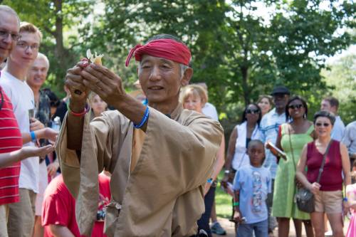 An elderly man in traditional attire performs for a diverse audience in a park, holding a small object with focus.
