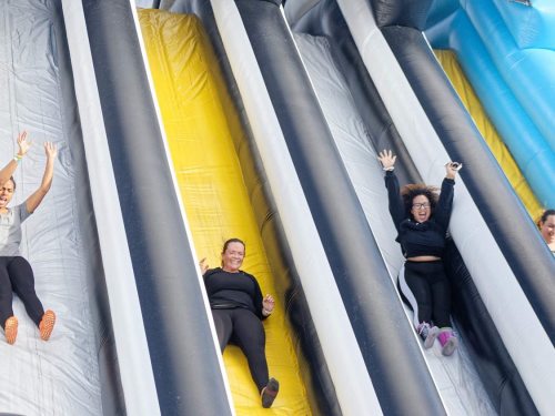 Three women joyfully sliding down colorful inflatable slides, arms raised in excitement.