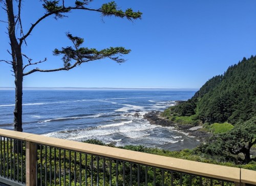 A scenic coastal view featuring waves, rocky shore, and lush green hills under a clear blue sky.