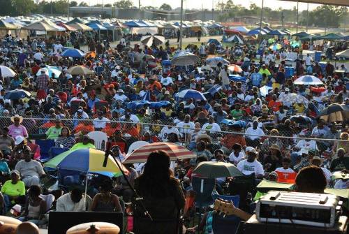 A large crowd of people sitting under umbrellas at an outdoor event, with tents and a stage in the background.