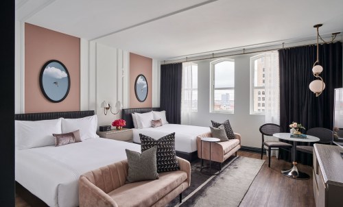 A stylish hotel room featuring two beds, modern decor, large windows, and a cozy seating area.