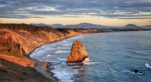 A scenic coastal view at sunset, featuring a rocky outcrop, gentle waves, and distant mountains under a cloudy sky.