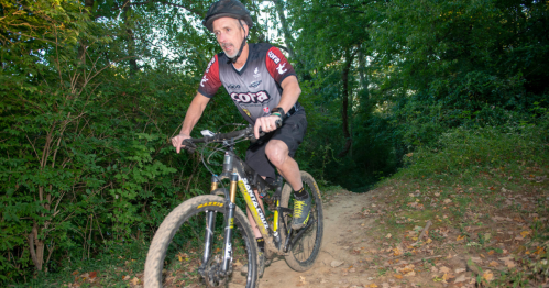 A cyclist rides a mountain bike on a dirt trail surrounded by greenery.