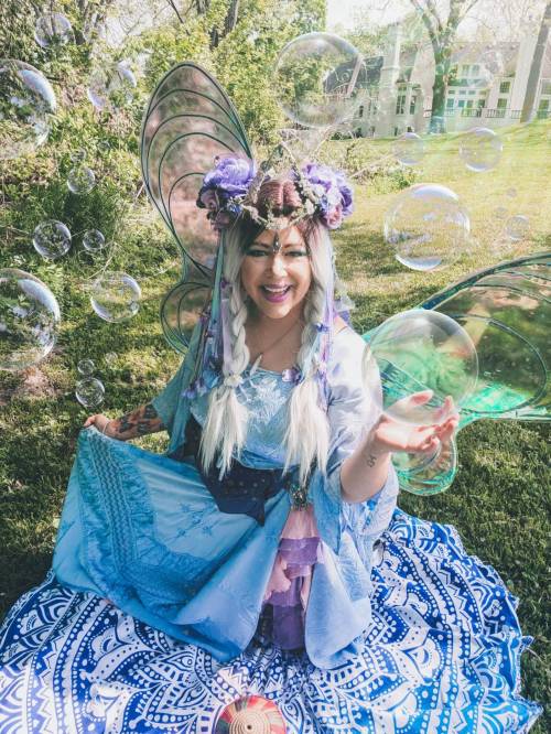 A smiling fairy with colorful wings sits on a patterned blanket, surrounded by floating bubbles in a sunny outdoor setting.