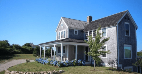 A charming gray shingle house with a porch, surrounded by green grass and blue hydrangeas under a clear blue sky.