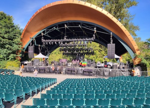 A large outdoor stage with a wooden canopy, surrounded by empty green seats and trees in the background.