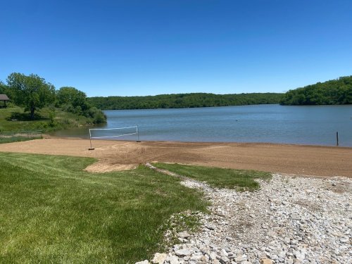 A sandy beach by a calm lake, with a volleyball net and green trees in the background under a clear blue sky.