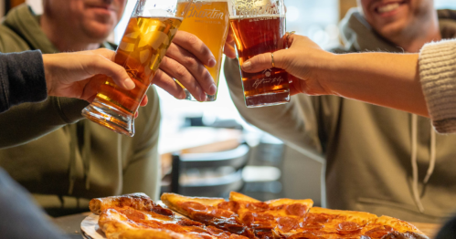 A group of friends toasting with beers over a pizza at a table.