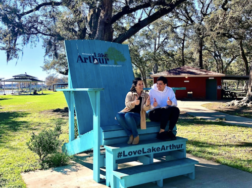 A couple sits on a large blue chair at Lake Arthur, smiling and giving thumbs up, with trees and a pavilion in the background.