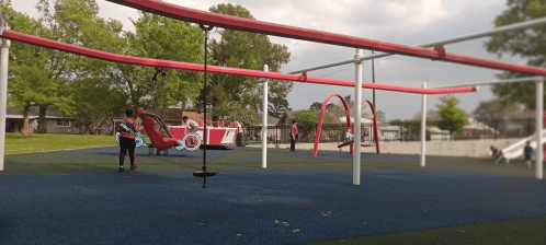 A playground with red swings and slides, children playing, and green trees in the background under a cloudy sky.