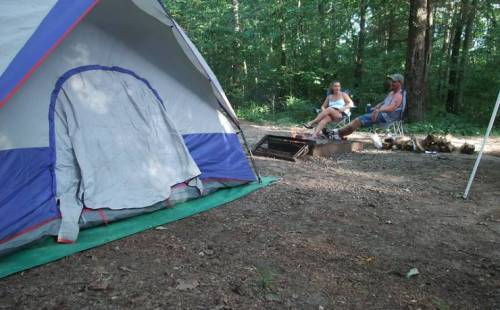 A tent in the foreground with two people sitting by a campfire in a wooded area.