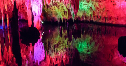 Colorful stalactites and stalagmites reflected in still water within a cave, illuminated by vibrant lights.