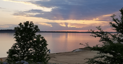 A serene lakeside view at sunset, with colorful clouds reflecting on the water and trees framing the scene.