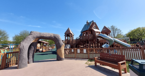 A playground featuring wooden structures, slides, and a large archway, set against a clear blue sky.