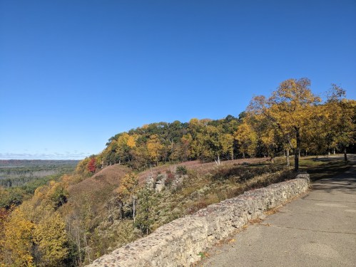 A scenic view of a hillside with autumn foliage under a clear blue sky, alongside a stone pathway.