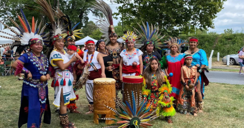 A group of people in vibrant traditional costumes, posing together with drums and feathered headdresses at an outdoor event.