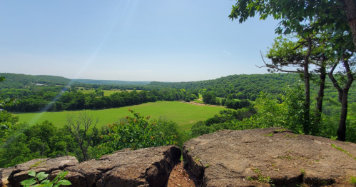 A scenic view from a rocky overlook, showcasing lush green fields and rolling hills under a clear blue sky.