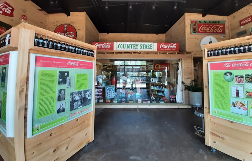 A rustic country store display featuring Coca-Cola memorabilia and products, with wooden structures and informative panels.