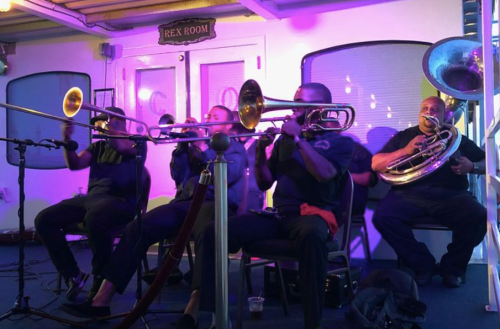A brass band performs on a boat, featuring musicians with trombones and a tuba, illuminated by colorful lights.
