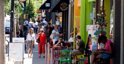A busy street scene with people walking, shops, and colorful outdoor seating on a sunny day.