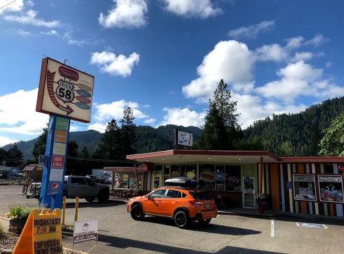 A colorful roadside diner with an orange car parked outside, surrounded by trees and mountains under a blue sky.