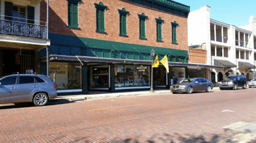 A street view of a brick road lined with shops, featuring green awnings and parked cars under a clear blue sky.