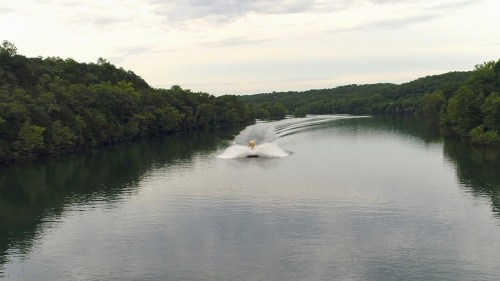 A person on a jet ski creates splashes while riding on a calm river surrounded by lush green trees.